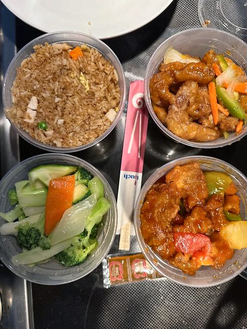 Four round plastic takeout containers on a steel counter, holding fried rice, mixed vegetables and two glazed dishes.