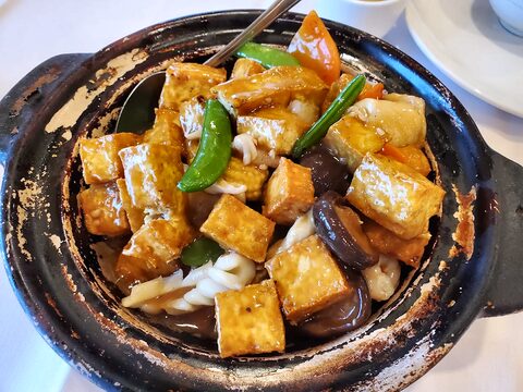 Glazed tofu cubes with mushrooms, snow peas and sliced vegetables filling a worn black clay pot on a white tablecloth.