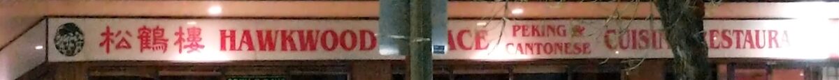 The illuminated white fascia at night, carrying red Chinese characters and red English lettering, partly crossed by a light pole and a bare tree.