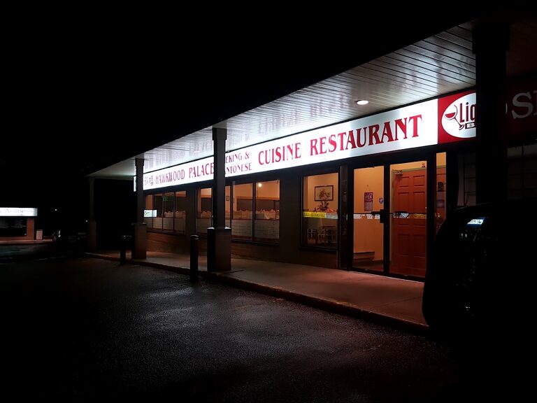 The storefront at night under the plaza’s covered walkway: the long lit fascia lettered in red above dark windows, and a warm-lit doorway at the end.