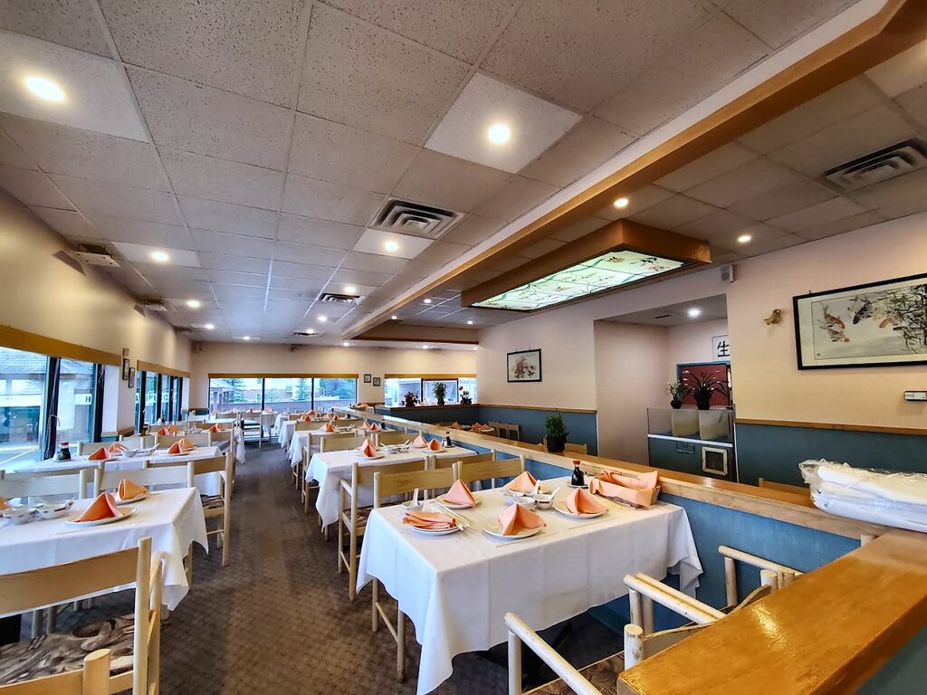 An empty, brightly lit dining room: white tablecloths with folded peach napkins, pale wood chairs, blue-green wainscoting, framed brush paintings and a backlit stained-glass ceiling panel.